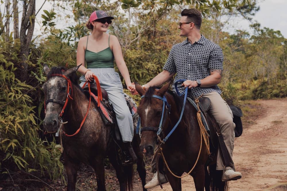 A smiling couple holds hands while riding horses on a forest trail.