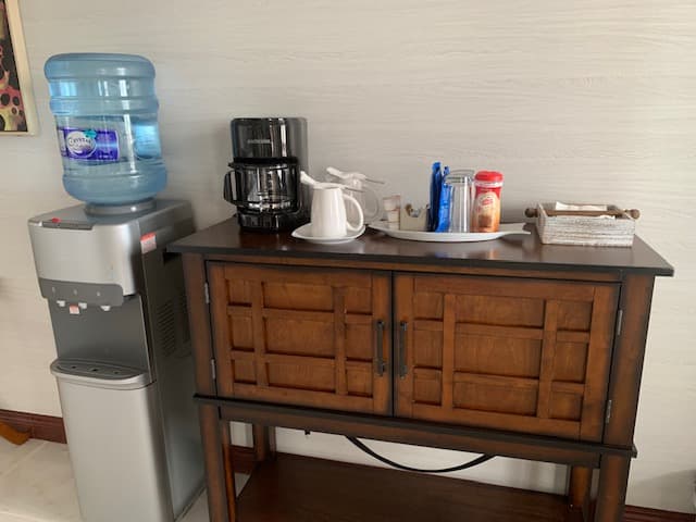A wooden cabinet holding a coffee maker, teapot, and condiments, next to a water cooler.