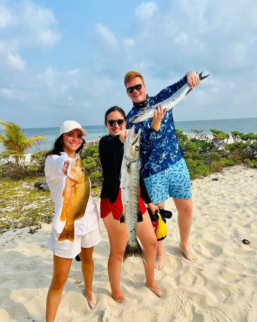 Three people stand on a beach holding large fish, with the ocean and palm trees in the background.
