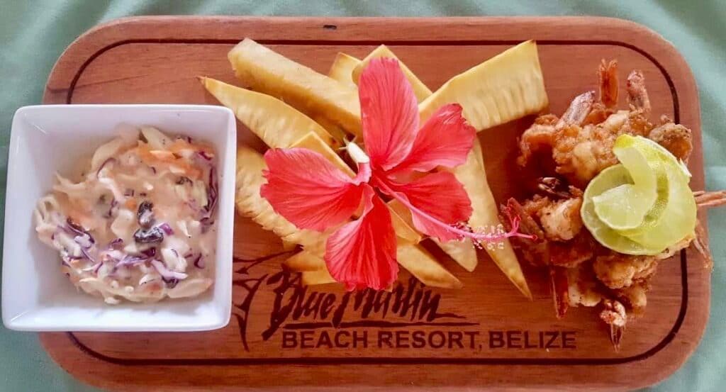 A wooden platter features coleslaw, fried shrimp with lime, and fried plantains, adorned with a red hibiscus flower.