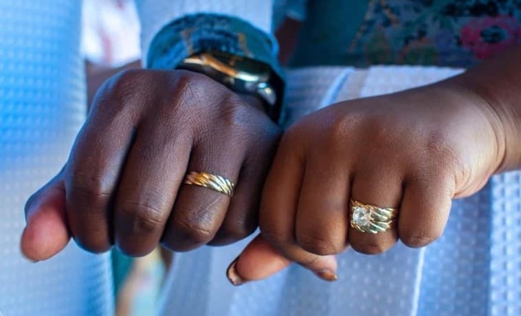 Two hands with wedding rings, one with a gold band and the other featuring a decorative ring, are clasped together.