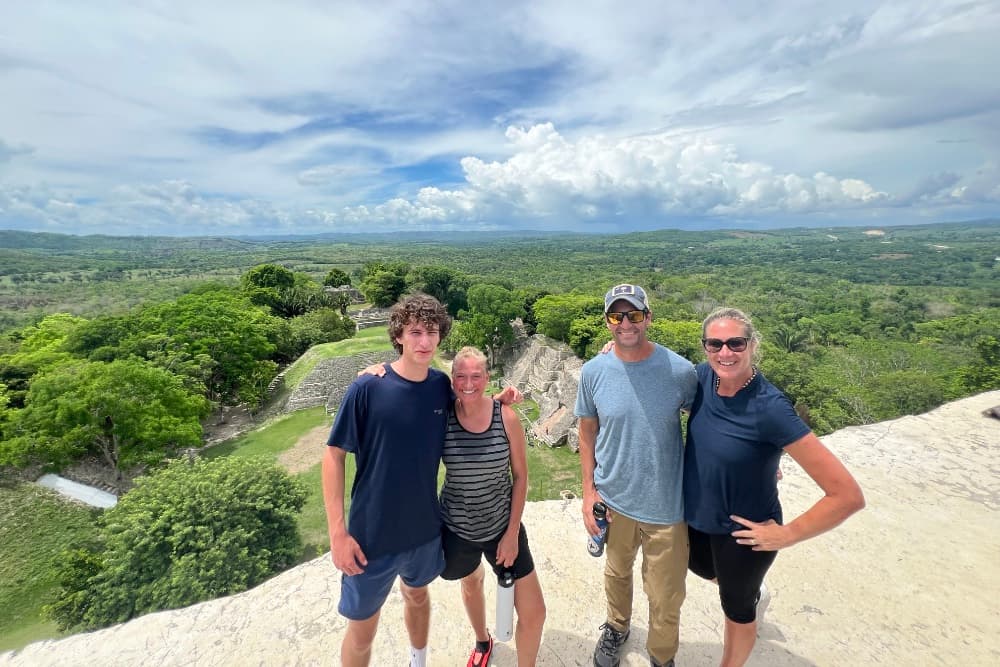 A group of four people stands on a scenic overlook with lush greenery and clouds in the background.