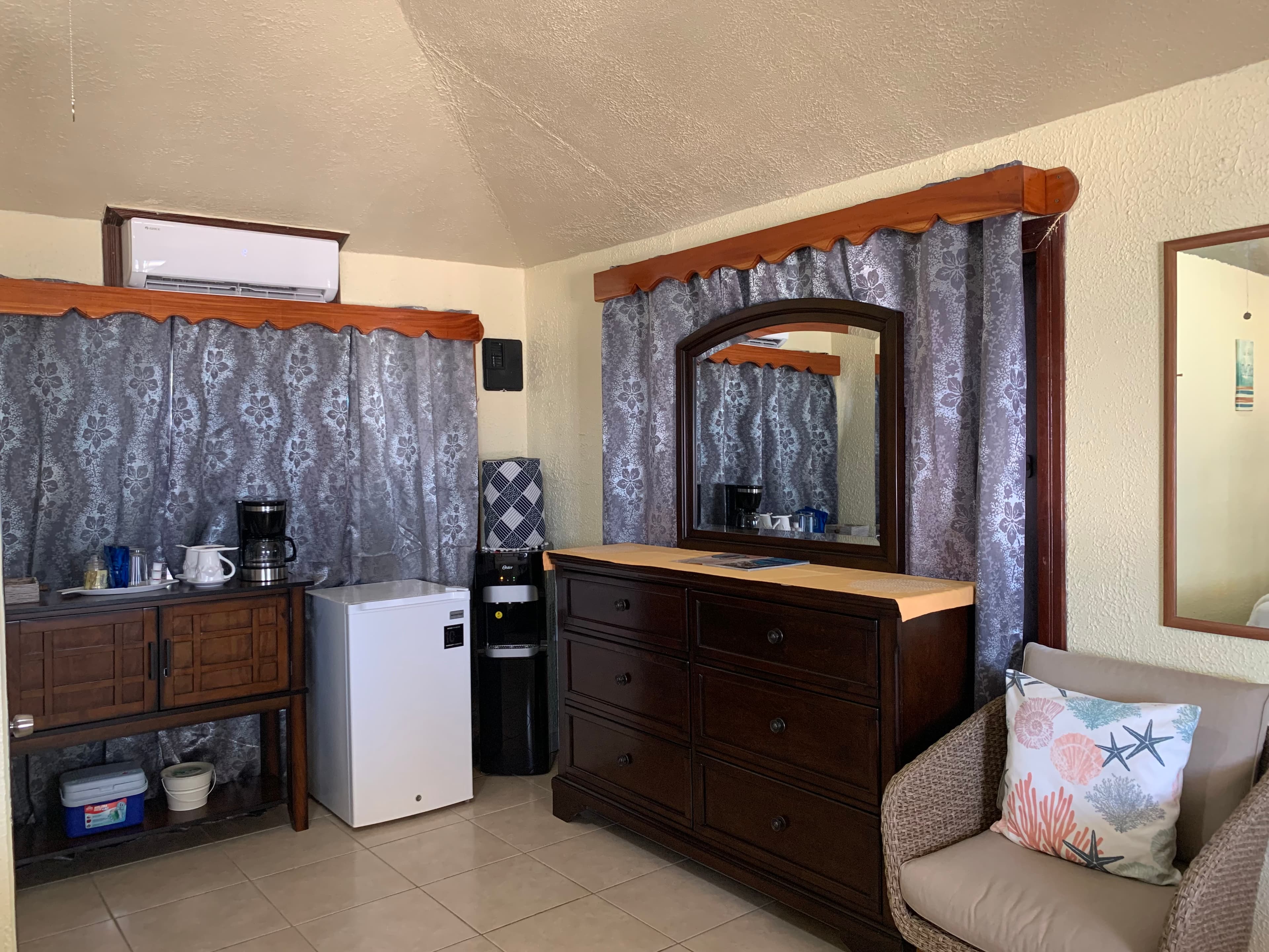 Interior of a small room featuring a dresser, mirror, mini fridge, coffee maker, and a cozy chair.