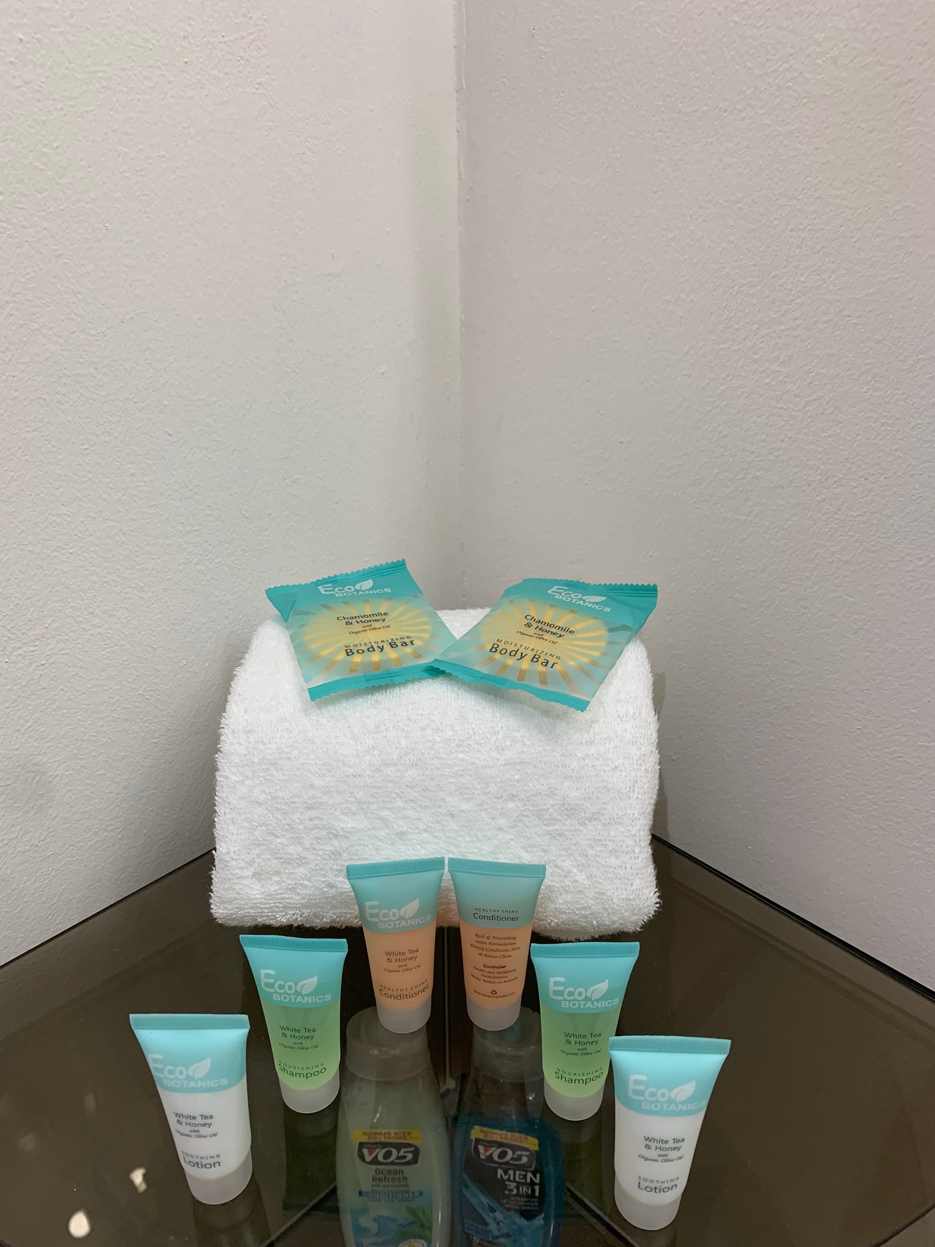 A neatly arranged display of various shampoo and conditioner travel-sized tubes on a glass surface, topped with a white towel and two packet samples.