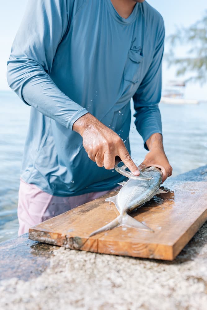 A person fillets a fish on a wooden cutting board by the water.