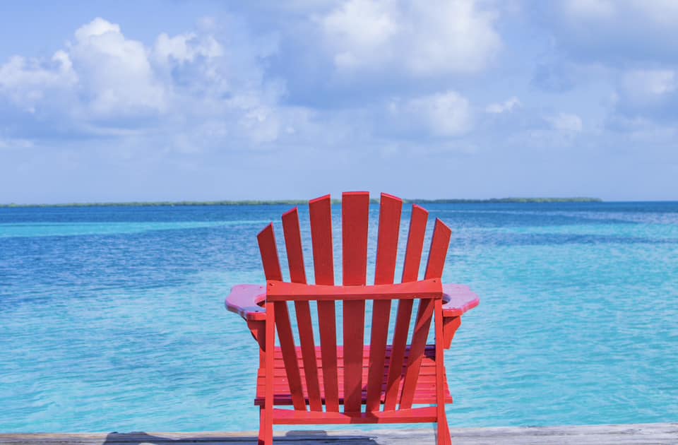 A red Adirondack chair faces a turquoise sea under a blue sky with fluffy clouds.