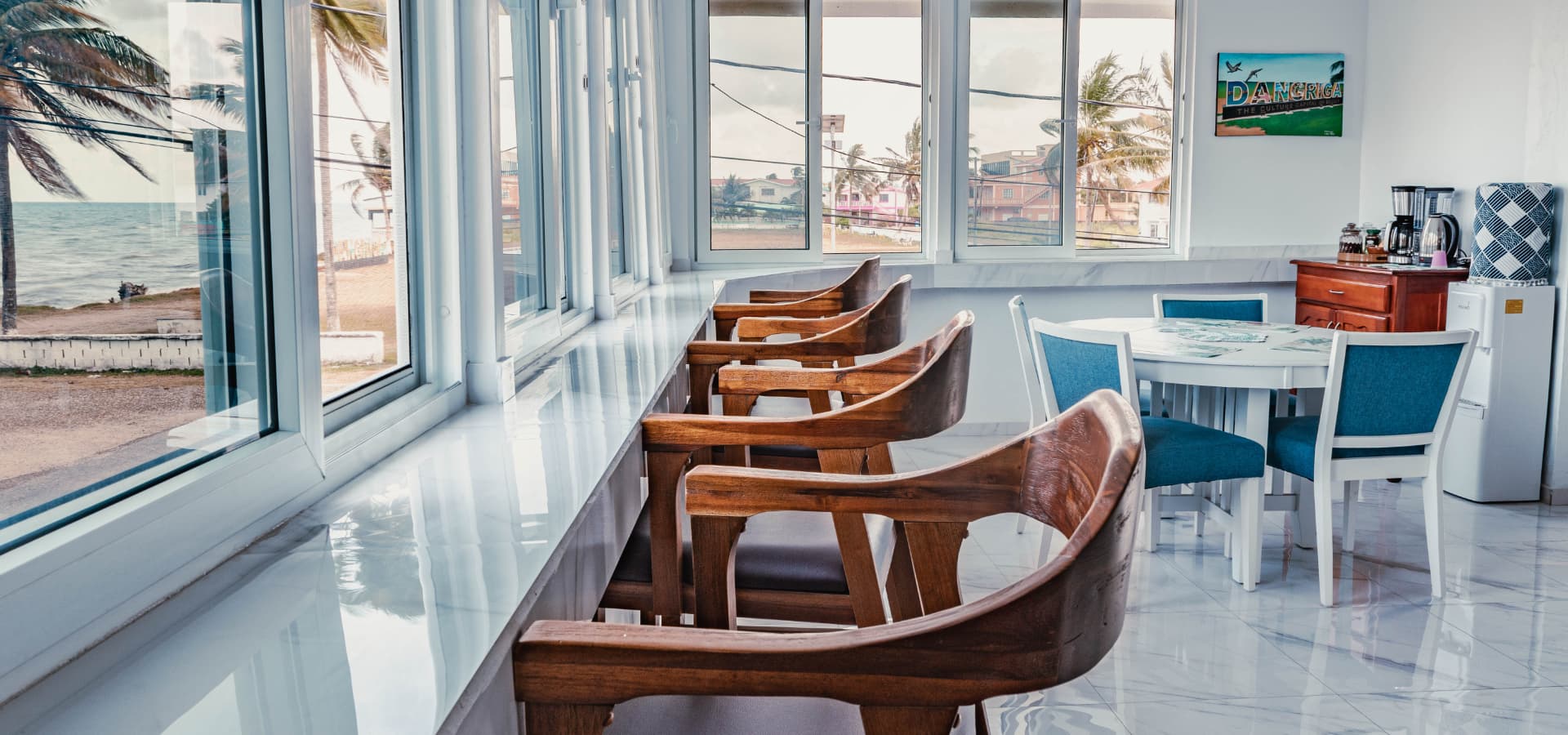A bright, modern dining area features wooden chairs and a view of palm trees and the ocean through large windows.