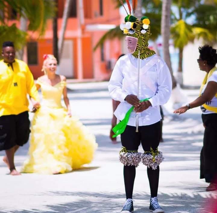 A person in a colorful costume stands on a beach pathway while a group of people walks in the background.