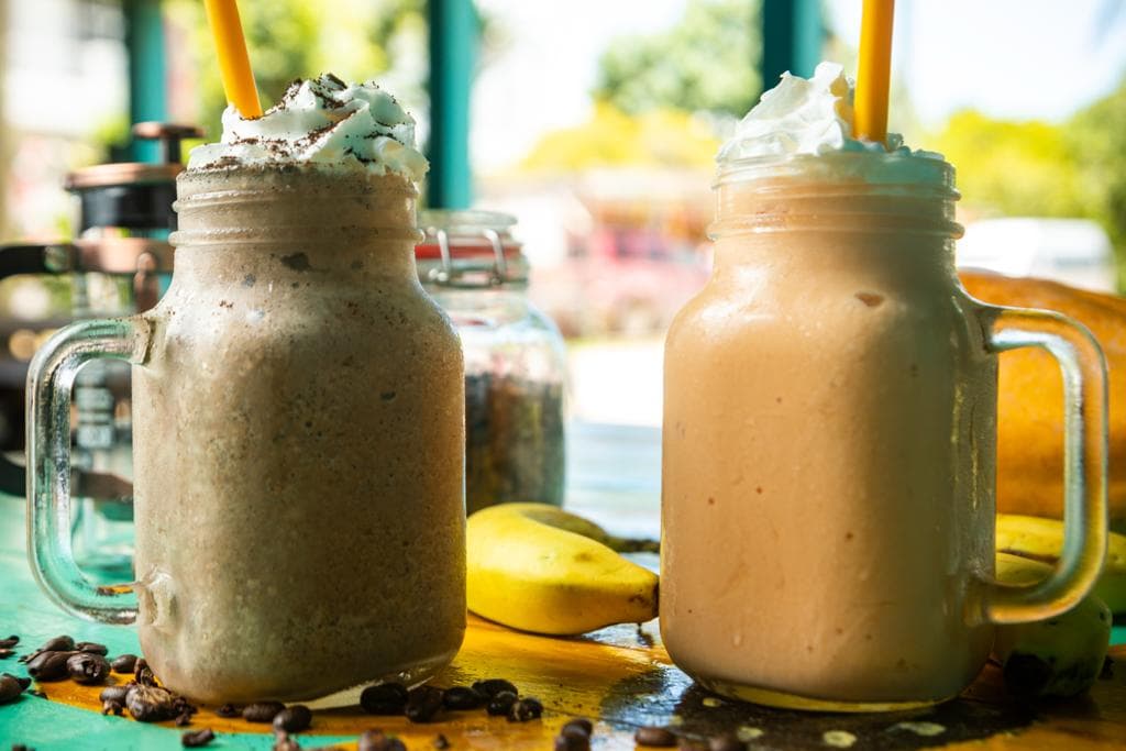 Two mason jars filled with smoothies, one chocolate with whipped cream and the other banana, on a colorful table.