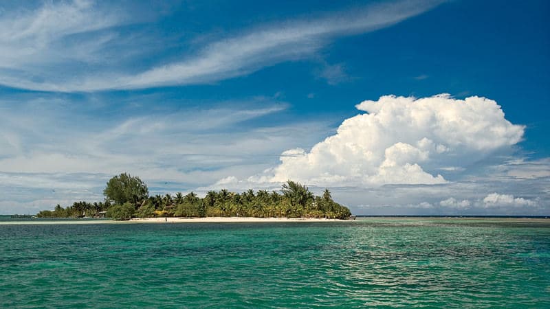 A tropical island surrounded by clear turquoise water under a blue sky with fluffy white clouds.