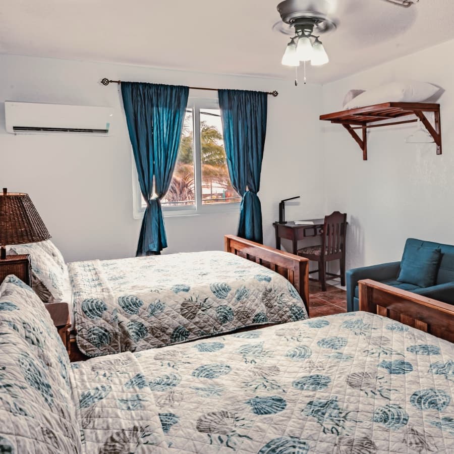 A bright bedroom featuring two quilted beds, a small desk, and a cozy seating area near a window.