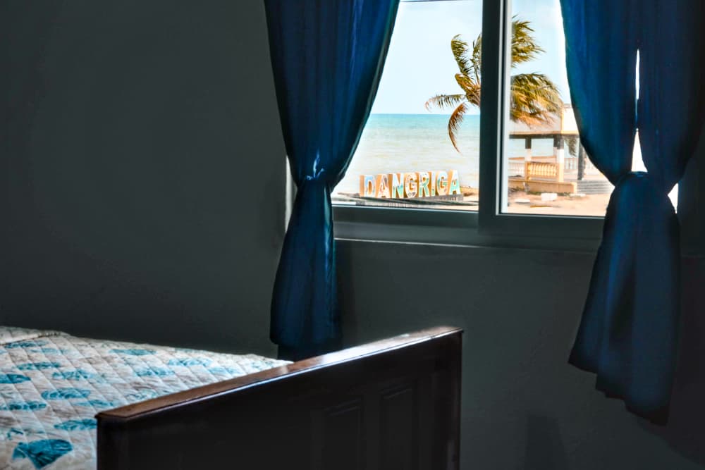 A view of a beach with palm trees and a sign reading "DANGRIGA" seen through a window from a room.