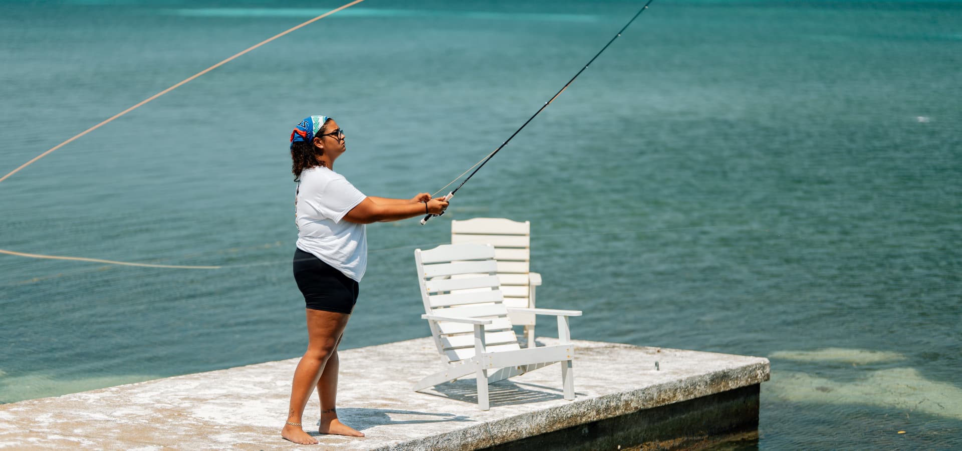A person fishes from a dock near calm waters with a white chair in the background.