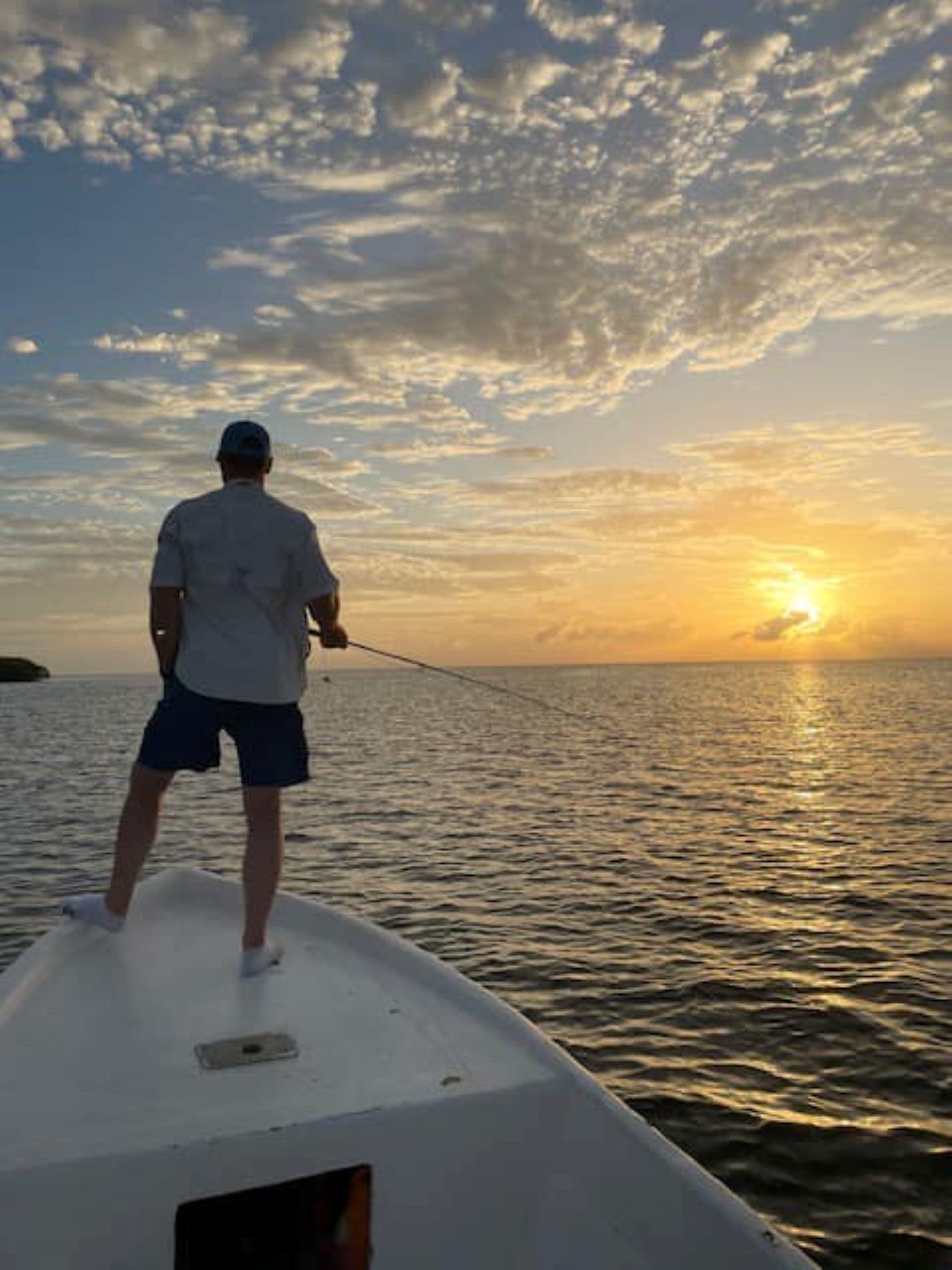 A person stands on the bow of a boat facing a vibrant sunset over calm waters.