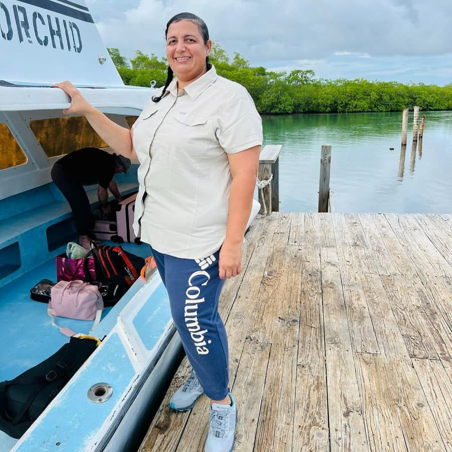 A person stands on a dock beside a boat, smiling, with bags in the background and lush greenery visible across the calm water.