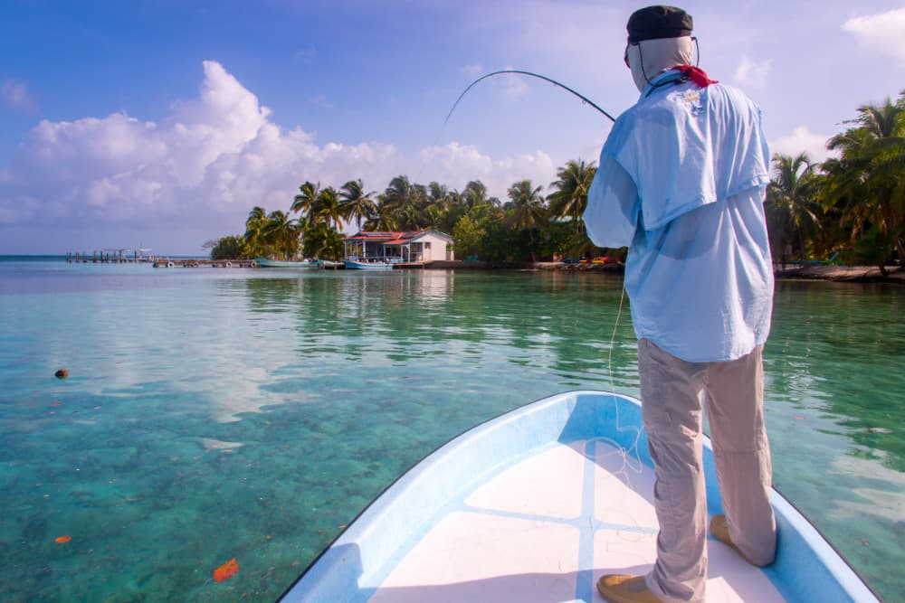 A man stands on a boat fishing in calm, clear waters surrounded by palm trees.