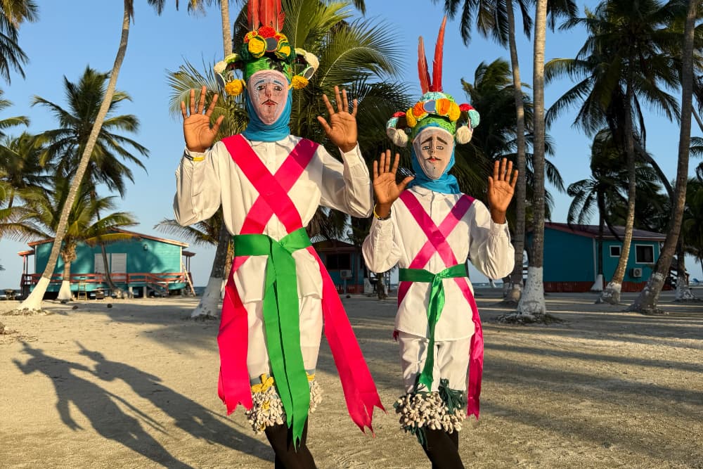 Two performers in colorful traditional attire pose with raised hands, surrounded by palm trees and beach houses.