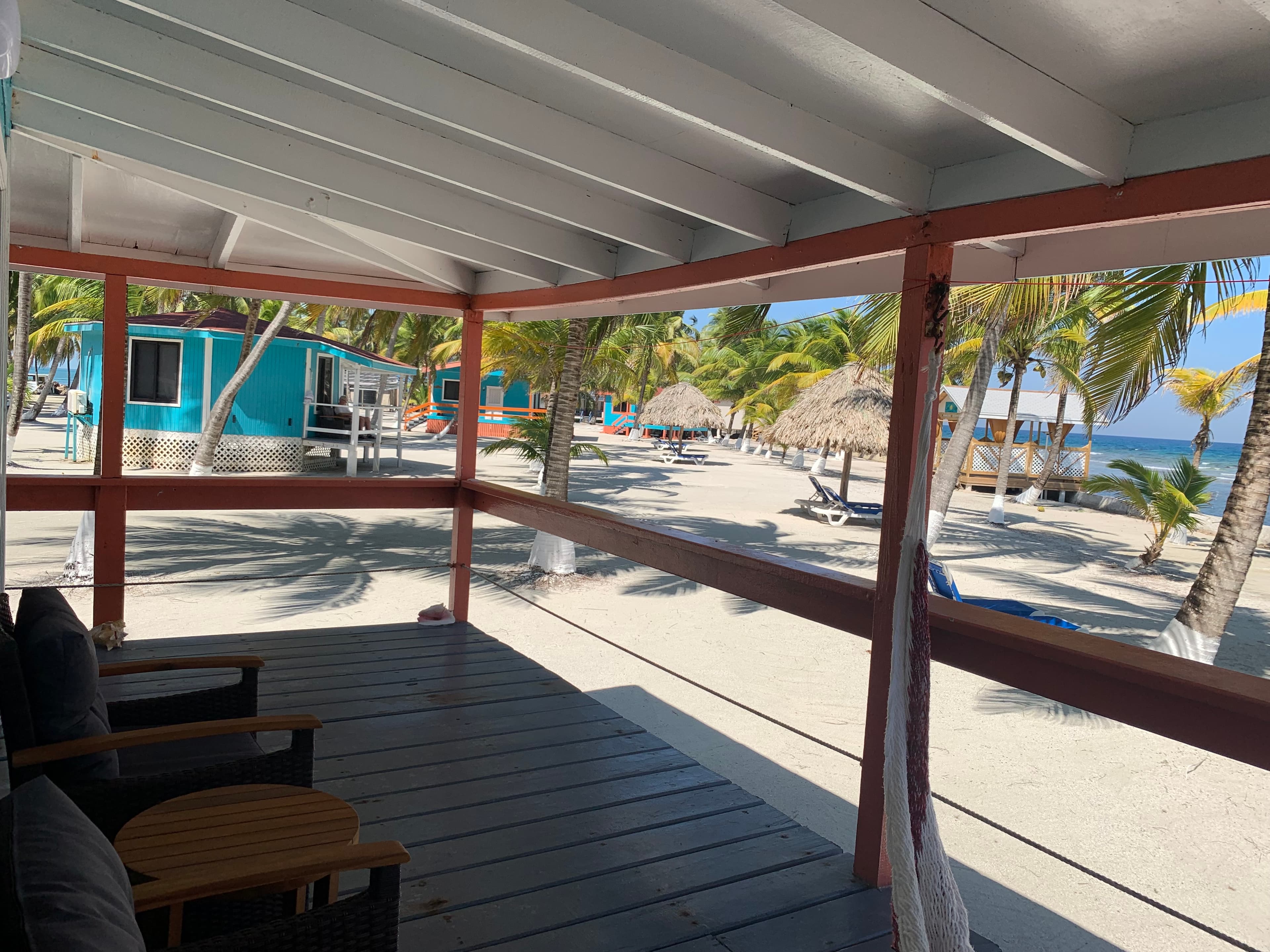 A view from a shaded porch overlooking a sandy beach with palm trees and colorful huts.