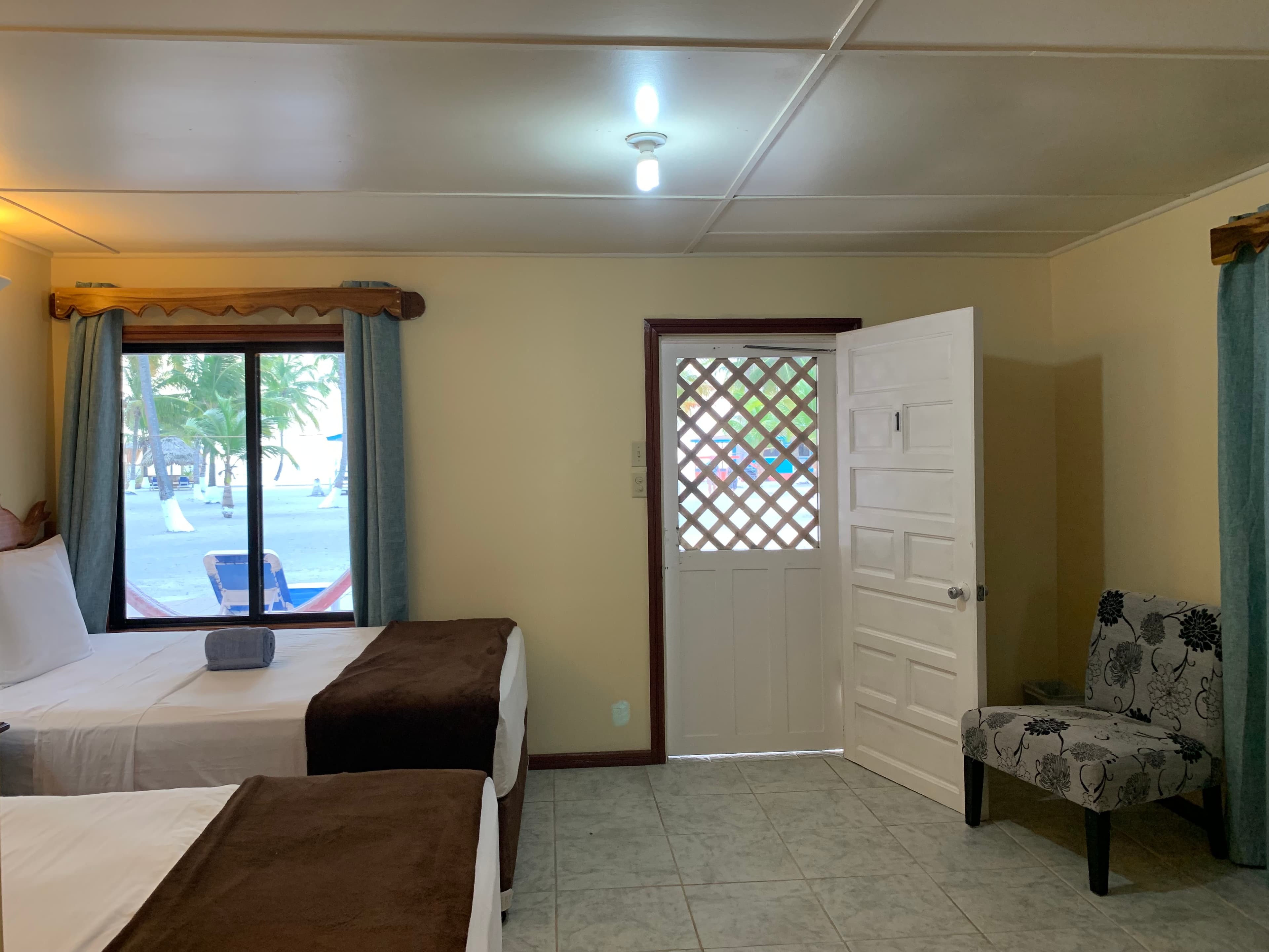 A brightly lit room with two beds, a chair, and an open door leading outside to a tropical view.