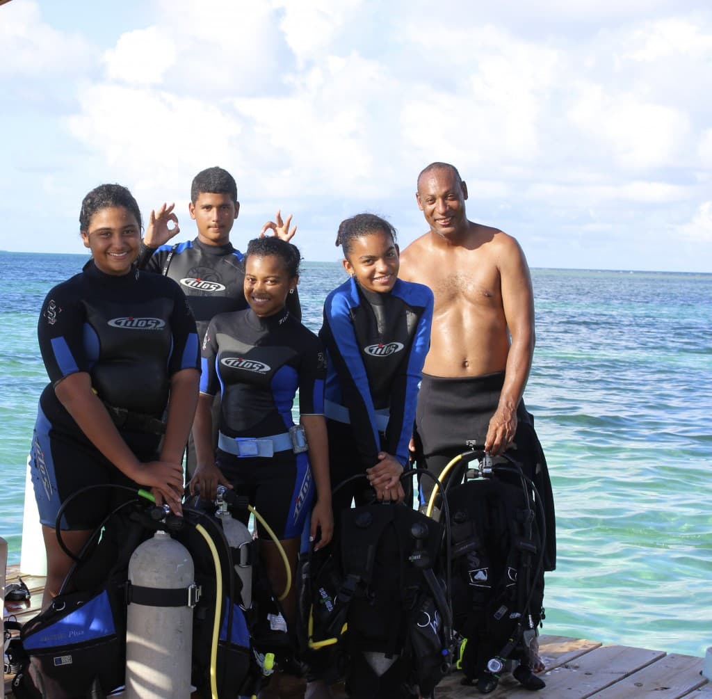 A group of five people in scuba gear poses by the water, smiling at the camera.