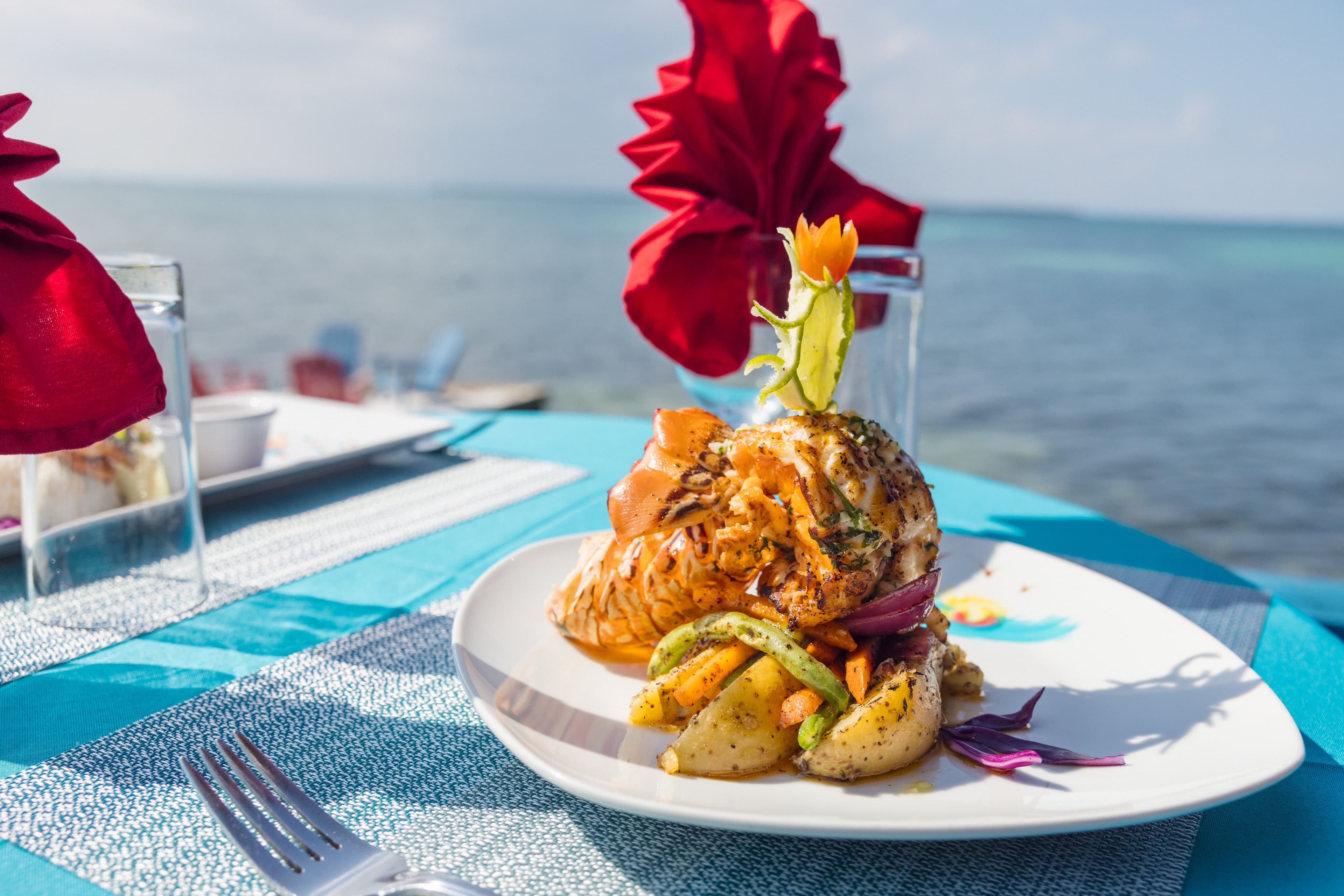A beautifully plated seafood dish with grilled lobster and colorful vegetables, set on a turquoise table by the water.