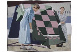 Two women in a quilted tapestry are lifting a large, patterned blanket together.