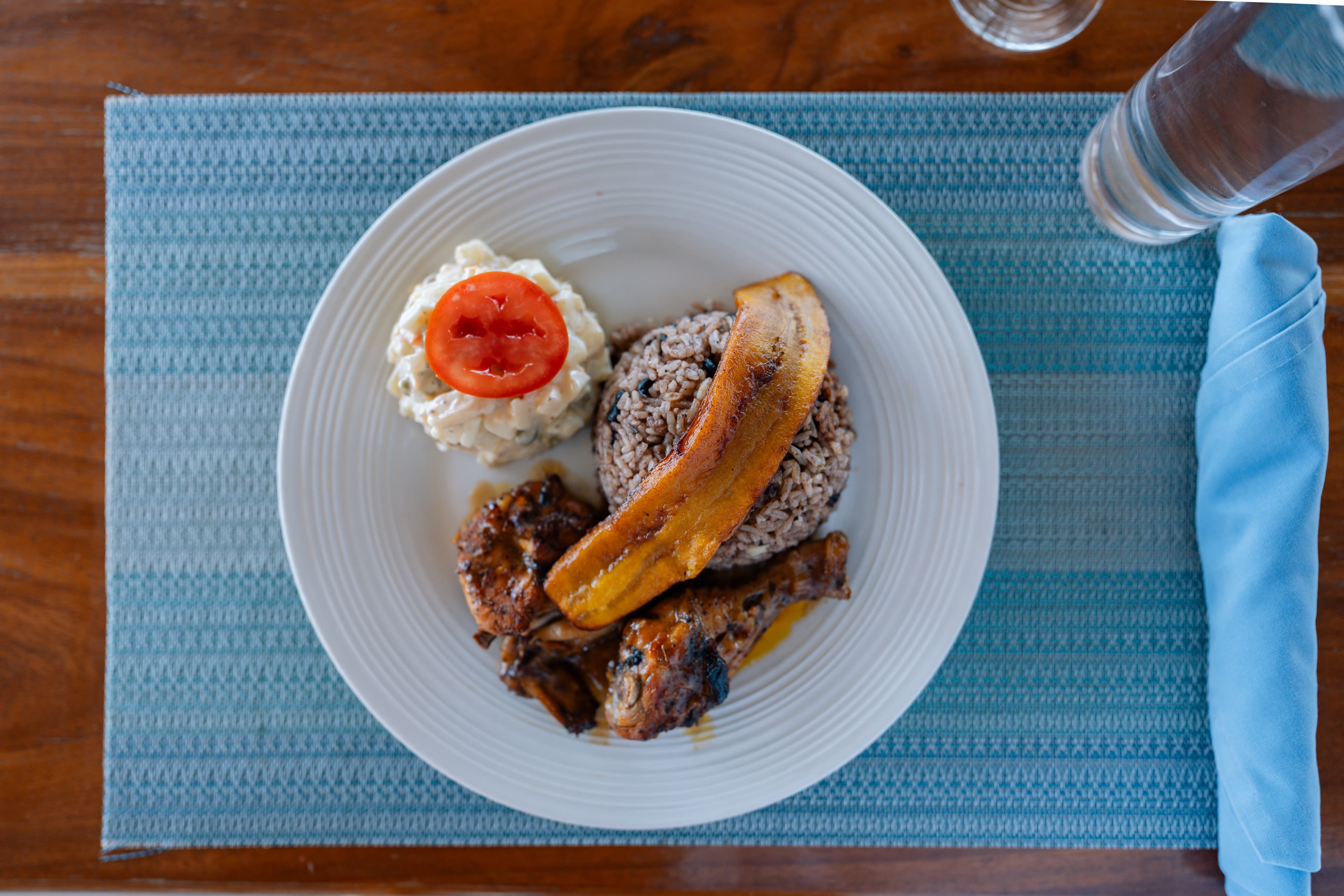 A plate featuring rice and beans, fried plantains, chicken, and a dollop of potato salad topped with a slice of tomato, arranged on a blue placemat.