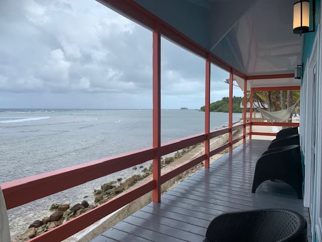 A covered balcony with black chairs overlooks a calm ocean and a distant shoreline under a cloudy sky.
