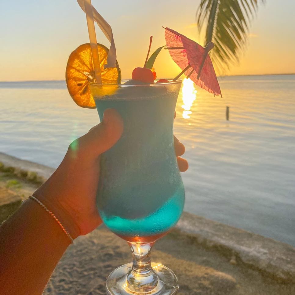 A hand holding a colorful blue cocktail with garnishes against a sunset over water.