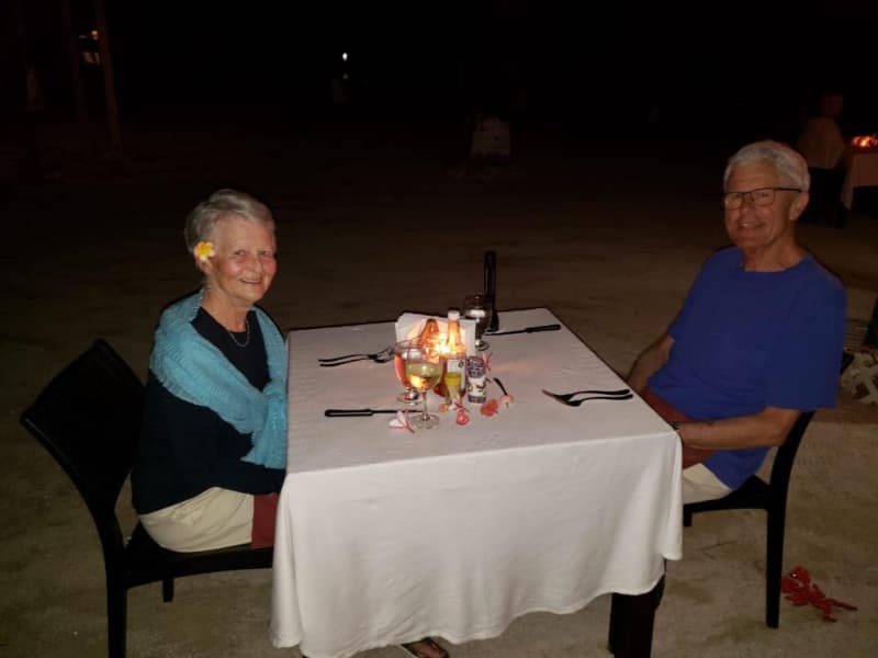 A smiling elderly couple sits at a candlelit dinner table on a beach at night. A smiling elderly couple sits at a candlelit dinner table on a beach at night.