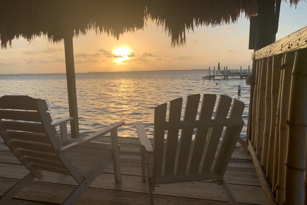 Two white lounge chairs on a pier overlooking a tranquil sunset on the water.