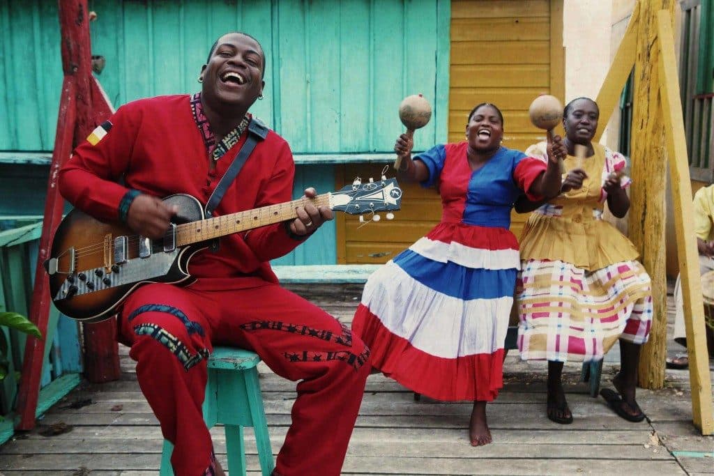 A musician laughs and plays guitar with two women in colorful dresses, celebrating together.