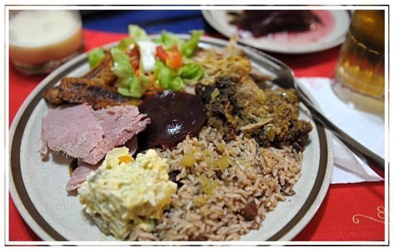A plate of assorted Caribbean food featuring rice, meat, salad, and a potato salad.