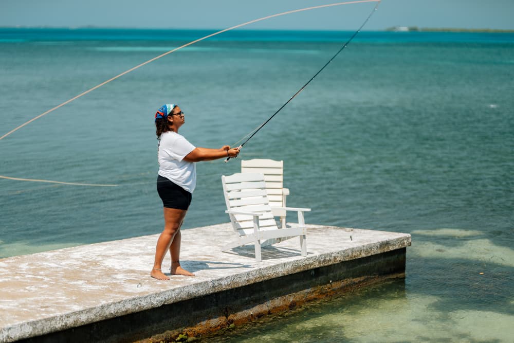 A person stands on a dock, casting a fishing line into clear blue water.