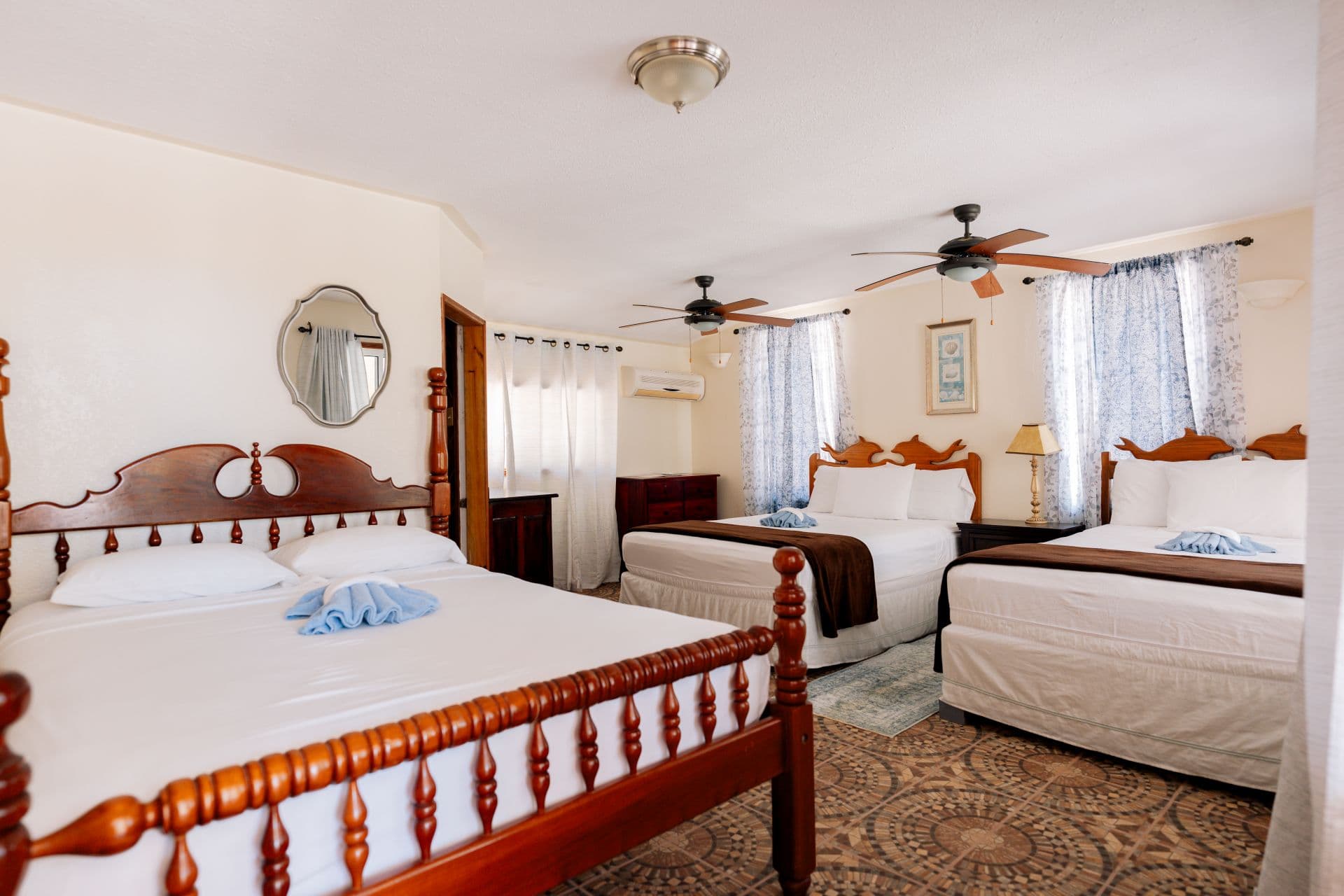 A spacious hotel room featuring three beds, two ceiling fans, and windows with light curtains.