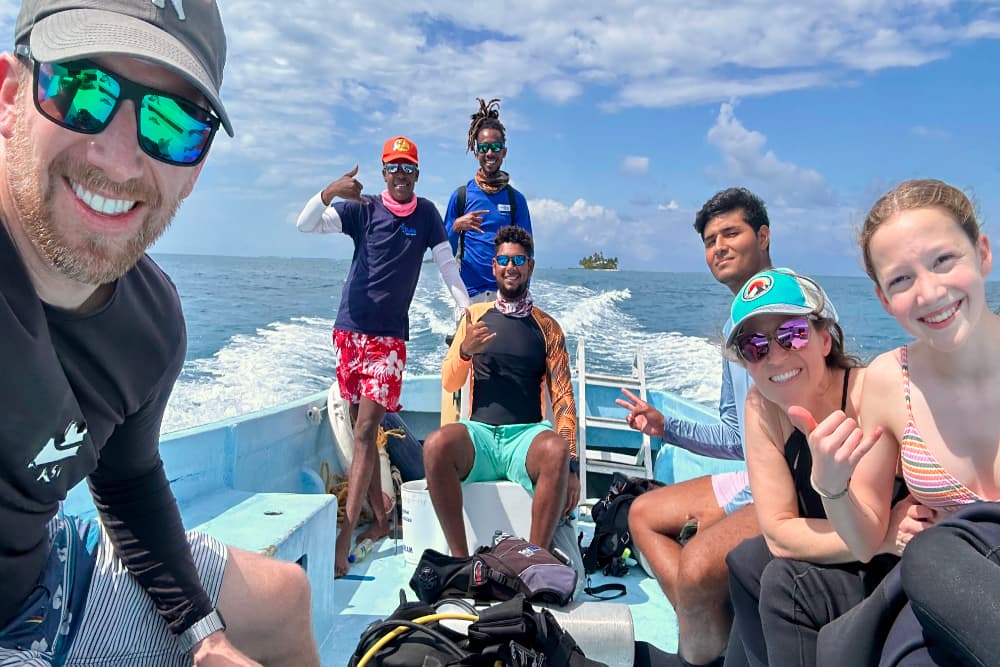 A diverse group of six people smiles and poses for a selfie while sitting in a boat on the water.