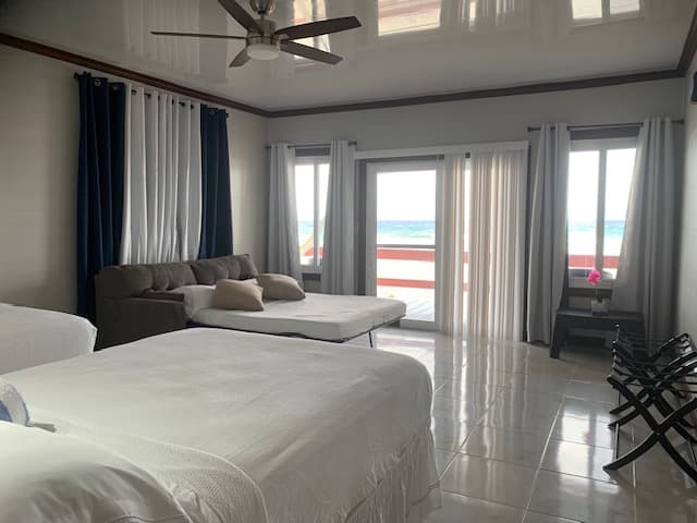 A bright, modern hotel room with two beds, a couch, and large windows overlooking a balcony and ocean view.