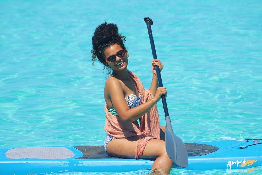 A woman smiles while sitting on a paddleboard in clear blue water.