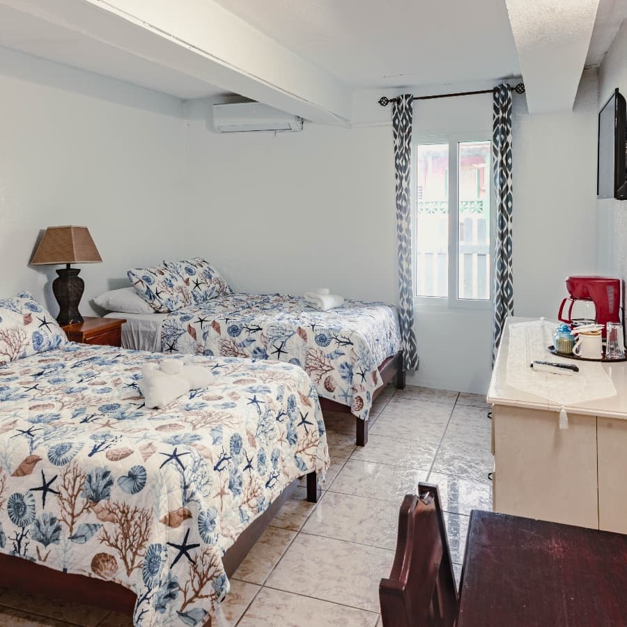 A cozy bedroom with two beds, decorated in a beach theme, featuring a window, lamp, and small table with coffee-making supplies.