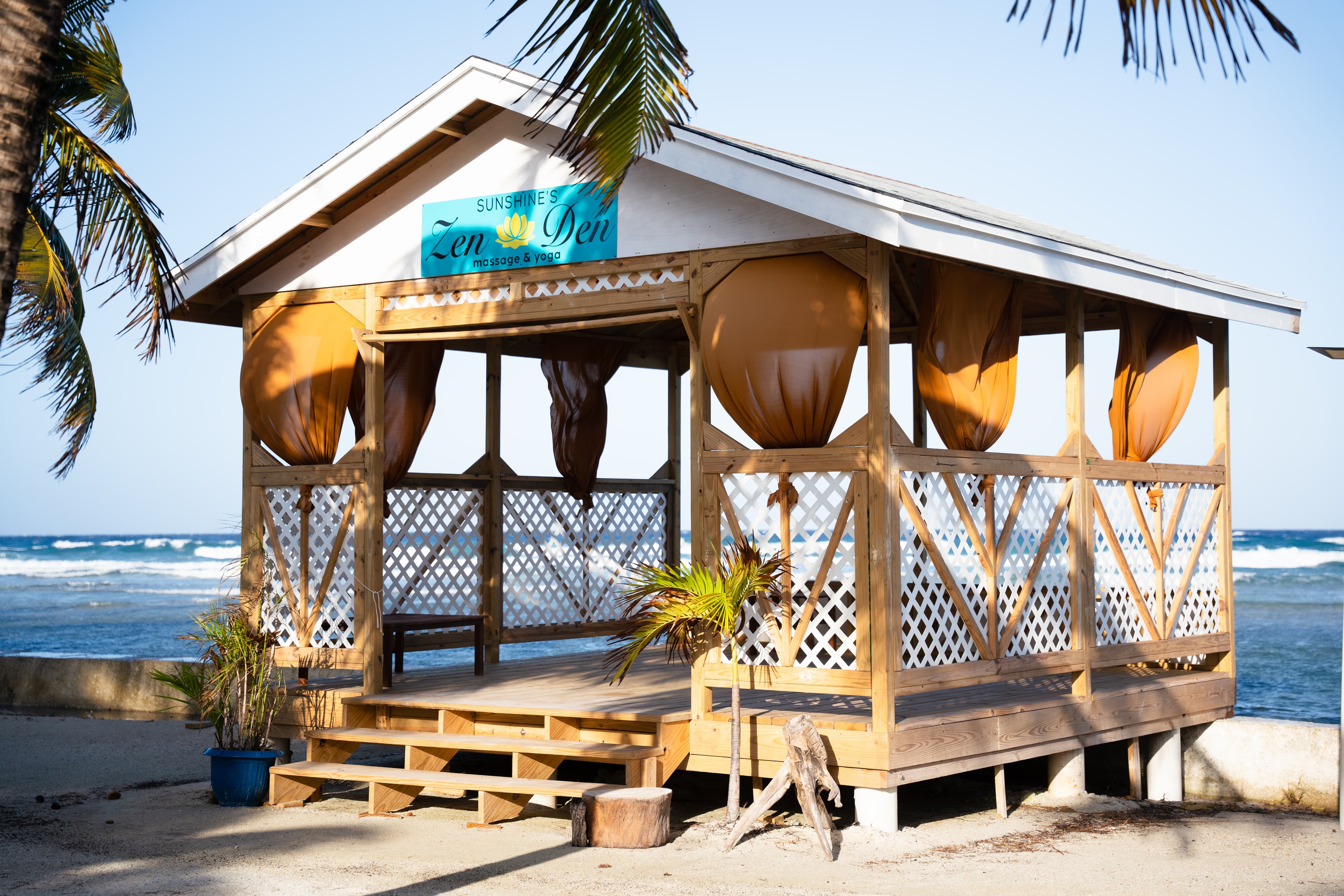 A wooden gazebo with orange curtains, labeled "Sunshine's Zen Den," overlooks a sandy beach and ocean waves.