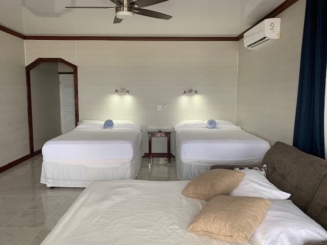 A bright hotel room featuring two double beds, a sofa, and a ceiling fan.