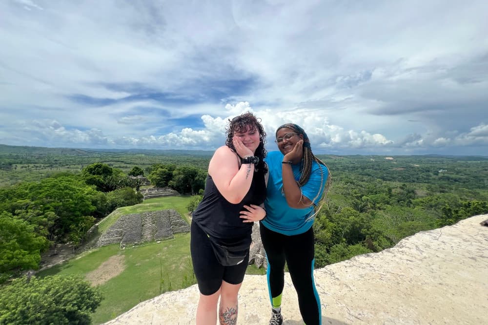 Two smiling women strike playful poses against a scenic landscape of greenery and clouds.