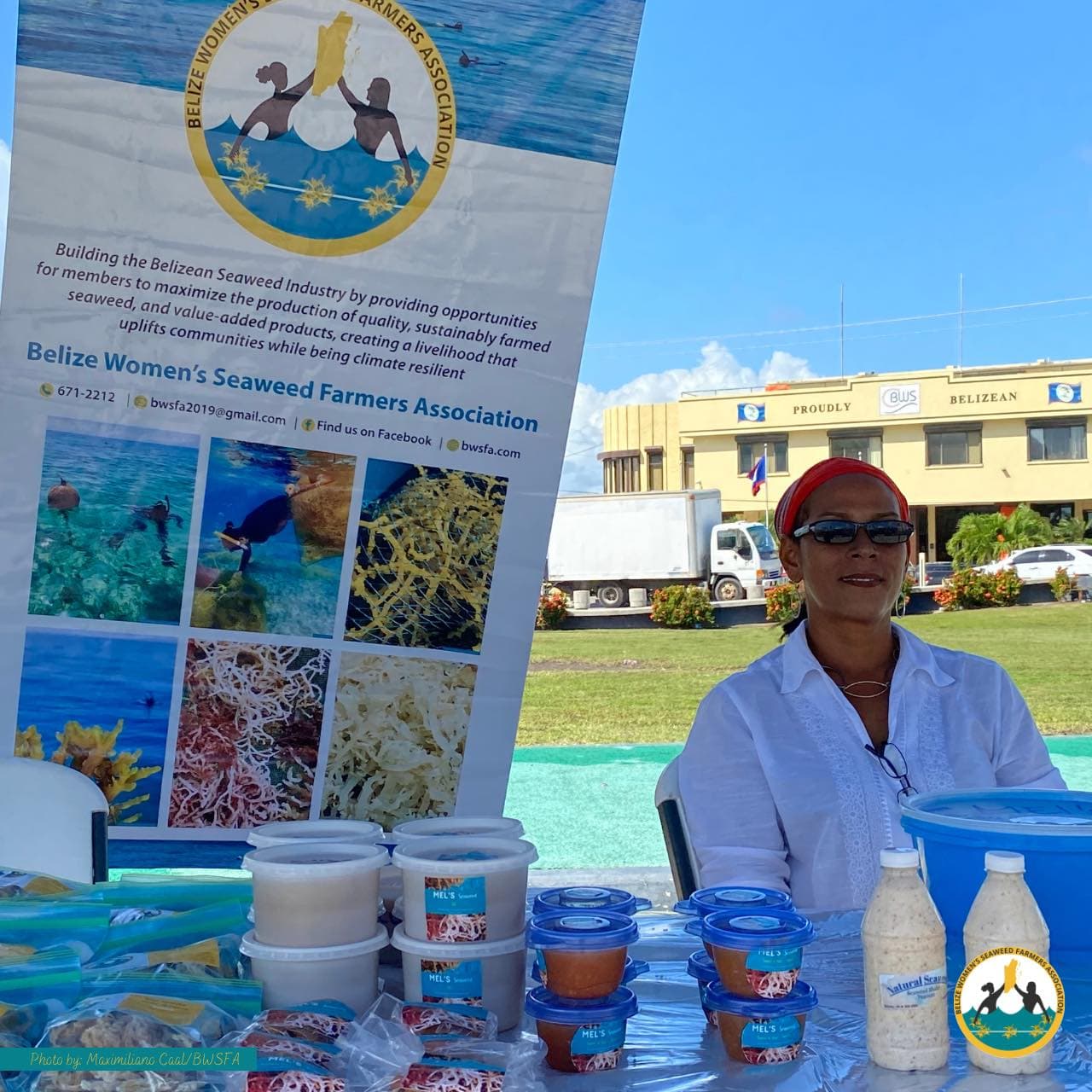 A woman sits at a booth promoting the Belize Women's Seaweed Farmers Association, surrounded by containers of seaweed products.