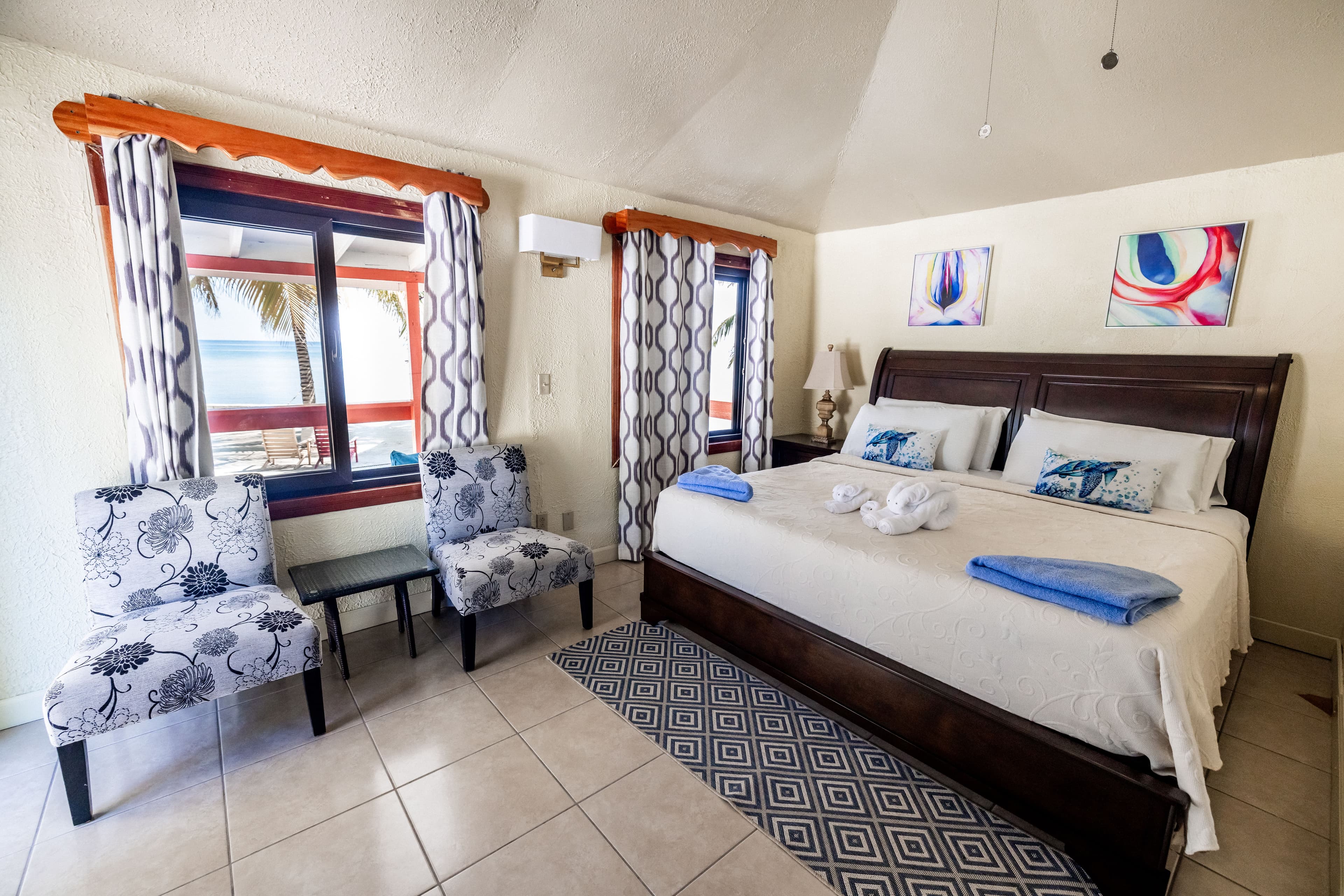A bright, airy bedroom featuring a large bed, two patterned chairs, and windows with scenic views.