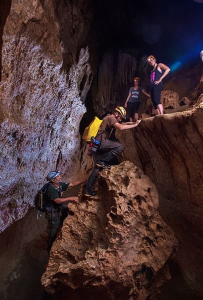A group of cavers explore a rocky cave, using headlamps and climbing gear.