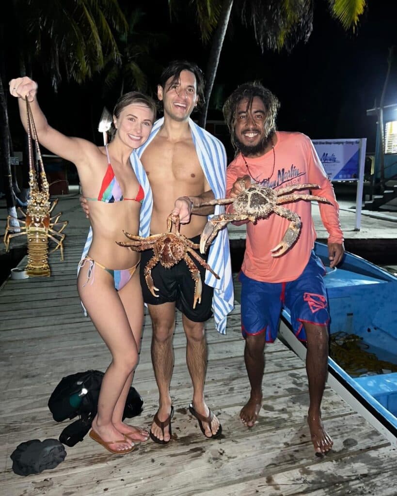 Three friends stand on a dock at night, each holding a large crab, with palm trees in the background.