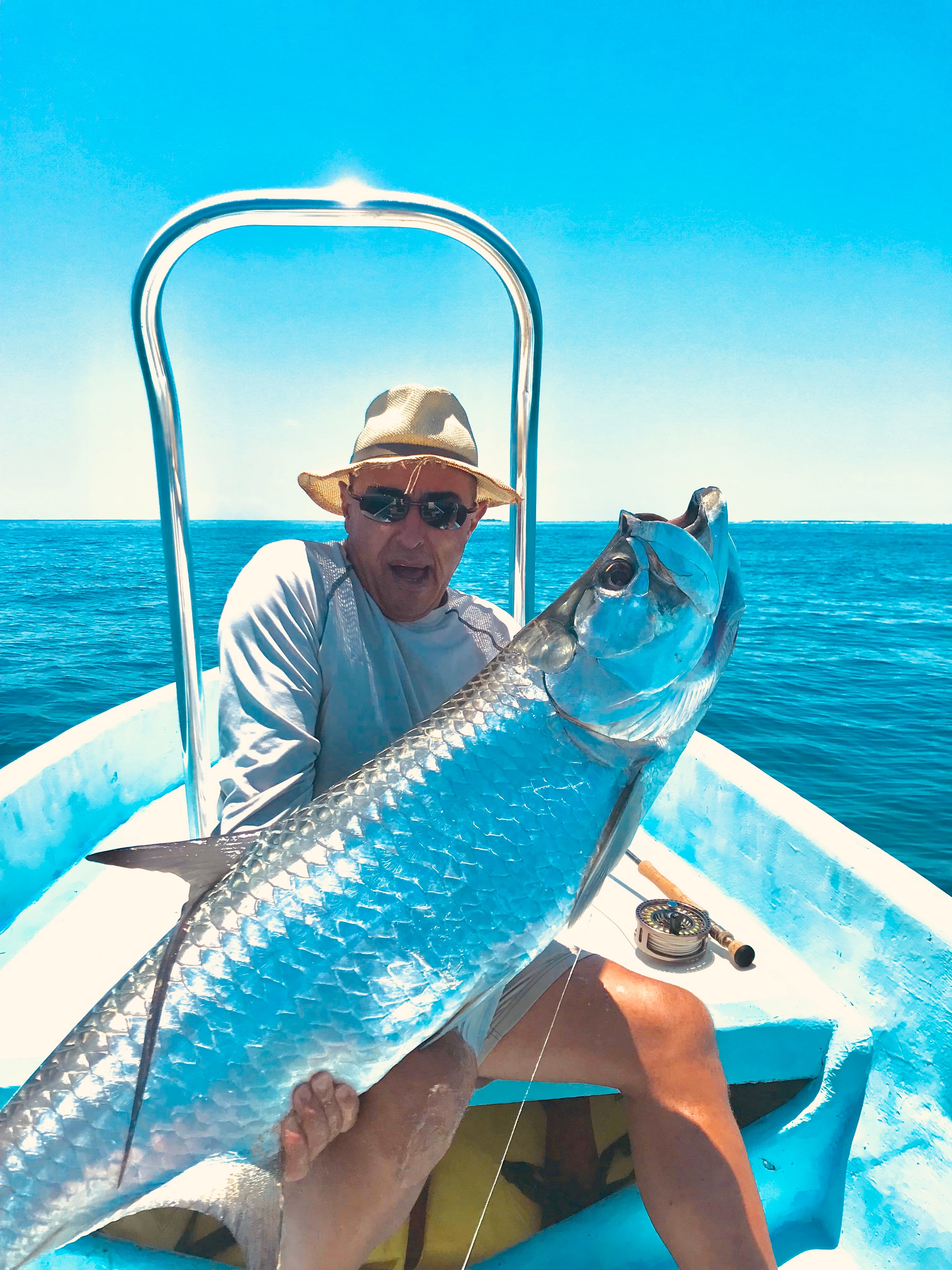 A man in a straw hat and sunglasses holds a large fish while sitting in a small boat on a sunny sea.