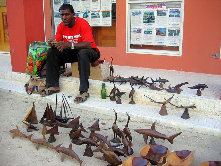 A man in a red shirt sits next to a variety of handmade wooden sculptures on a sidewalk.