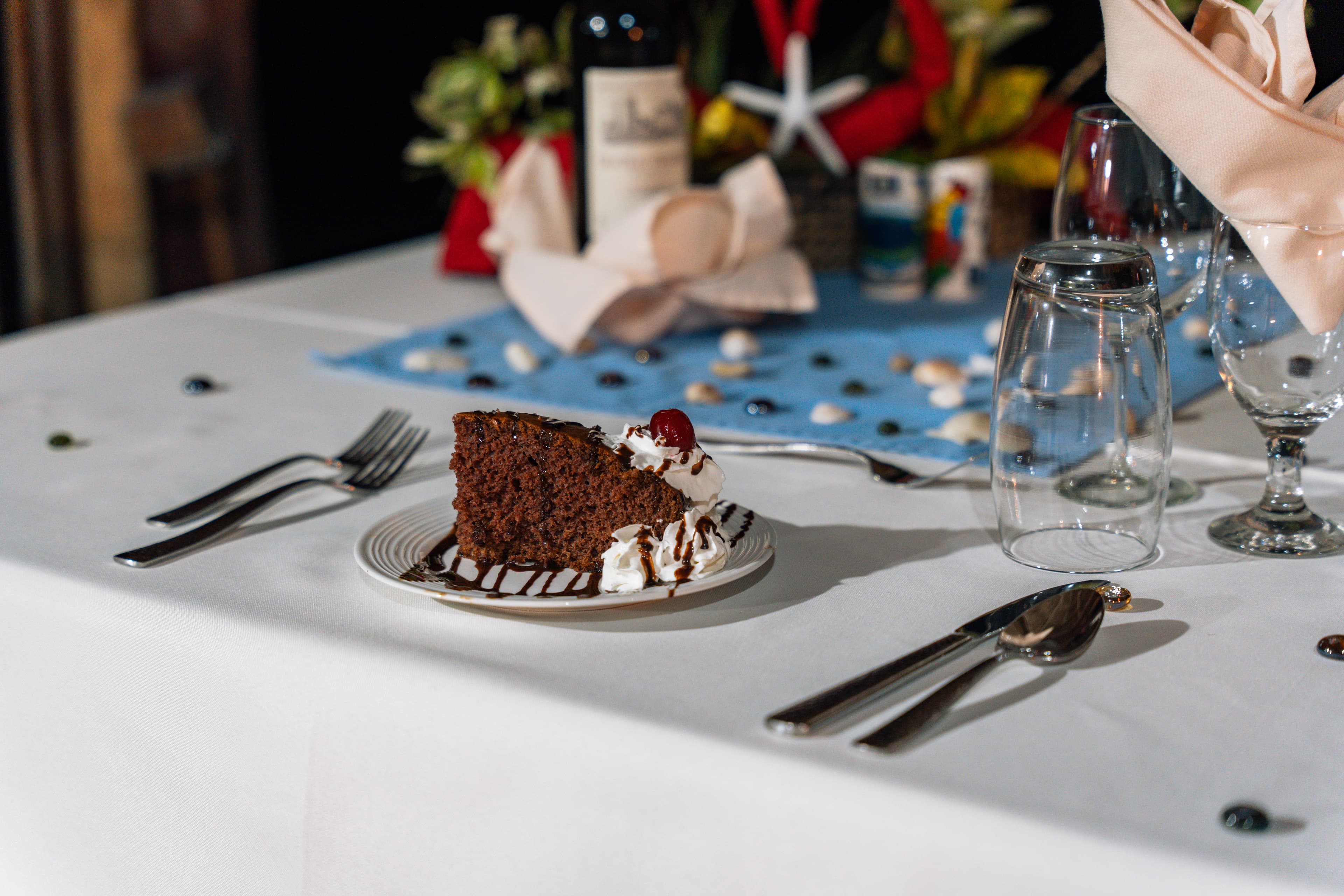 A slice of chocolate cake topped with whipped cream and a cherry sits on a table set for dining.
