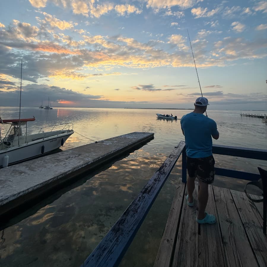 A person fishes off a wooden dock at sunset, with boats visible in the calm water.